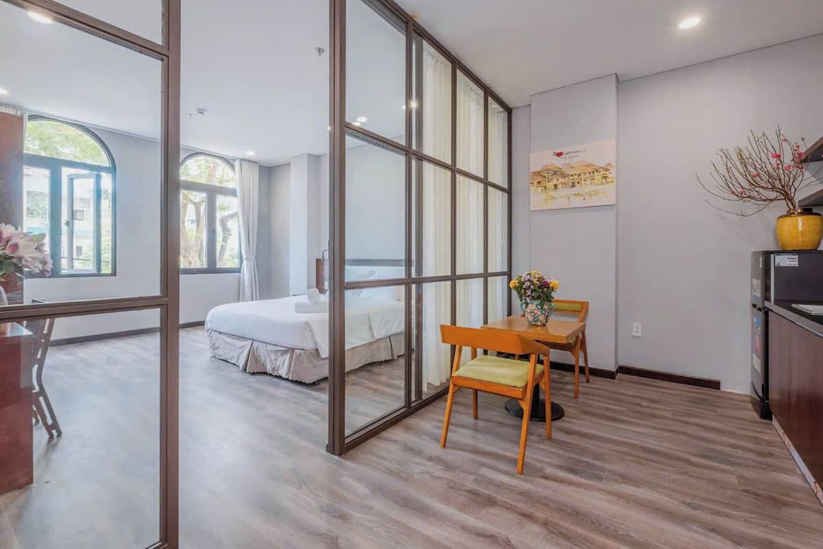 A modern hotel room with a large sliding glass door leading to a balcony, a bed, a dining table with chairs, and a kitchen area.