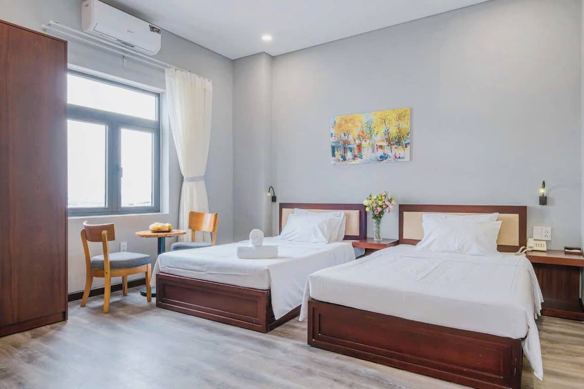 A hotel room with two beds, a desk, a chair, a window with curtains, and a painting on the wall.