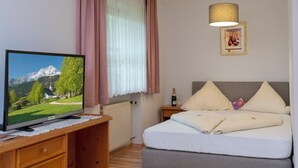 A bedroom with a bed, a TV on a wooden desk, a nightstand with a bottle and glasses, and a painting on the wall. - Apartment type 4 (Anger)