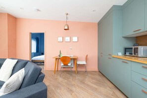 A modern hotel room with a sofa, a dining table with chairs, a kitchenette, and a bed with a blue wall behind it.