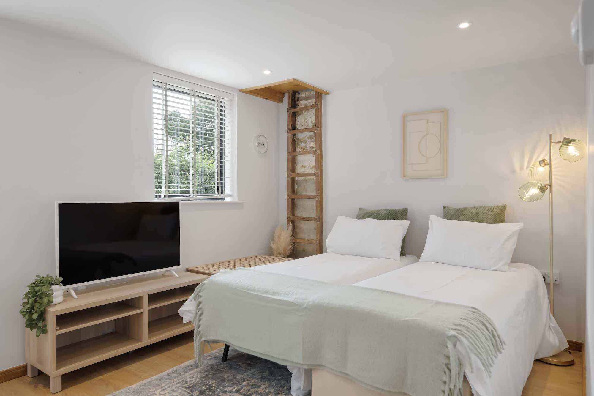 A bedroom with a bed, a TV on a wooden stand, a window with blinds, a ladder, a wall-mounted light fixture, and a small plant.
