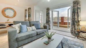 A modern living room with a grey sofa, a coffee table with a plant, and a large window with curtains. - Staithes House (Gateshead)