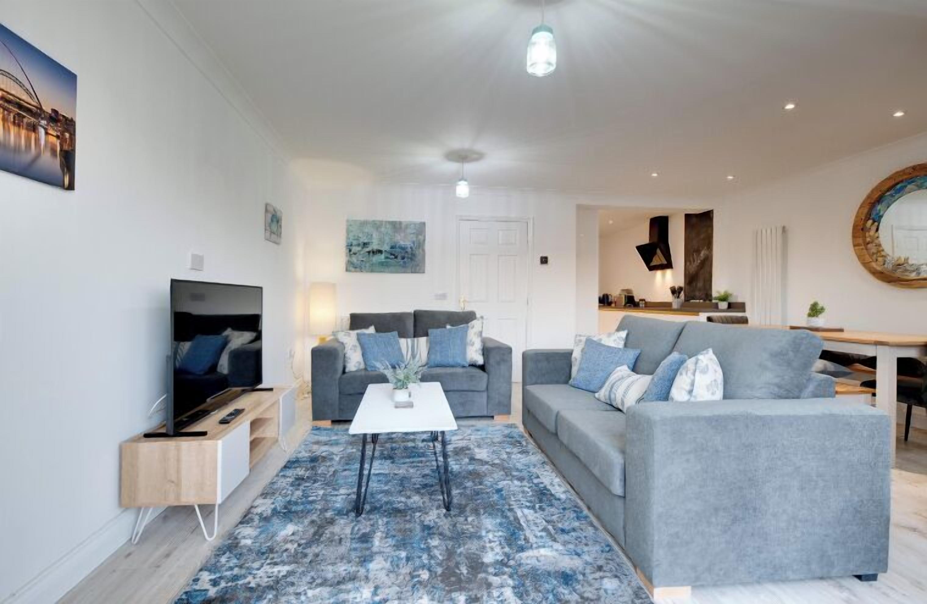A modern living room with a flat-screen TV, two grey sofas, a coffee table, and a blue patterned rug.