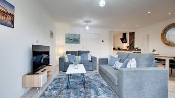 A modern living room with a flat-screen TV, two grey sofas, a coffee table, and a blue patterned rug.