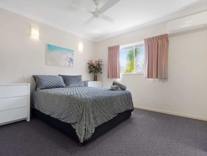 Ein Schlafzimmer mit Bett, Nachttisch, Deckenventilator, Fenster mit Vorhängen und einem Wandbild.
