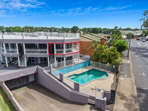 A multi-story building with a rooftop terrace, a swimming pool, and a street view with cars and greenery.