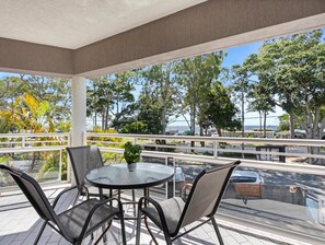 Ein überdachter Außenbereich mit rundem Tisch und Stühlen, einem Blumentopf und Blick auf Meer und Bäume.
