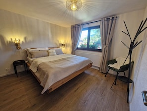 A bedroom with a wooden bed, two bedside tables, a window with curtains, and a chair. - Black Forest Queen House (Bühlertal)