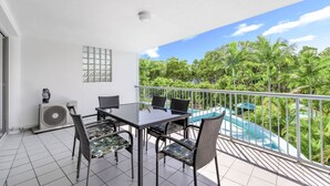 A balcony with a glass table and chairs, a white ceiling, and a view of a pool and tropical trees. - Hervey Bay Resort Stay with Balcony & Parking (Torquay)