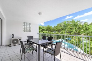 Un balcon avec une table et des chaises en verre, un plafond blanc et une vue sur une piscine et des arbres tropicaux.