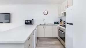 A modern kitchen with white cabinetry, a microwave, a sink, and a clock on the wall. - Hervey Bay Resort Stay with Balcony & Parking (Torquay)