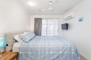 Une chambre à coucher avec un lit, un ventilateur de plafond, un téléviseur fixé au mur et une fenêtre avec des rideaux.