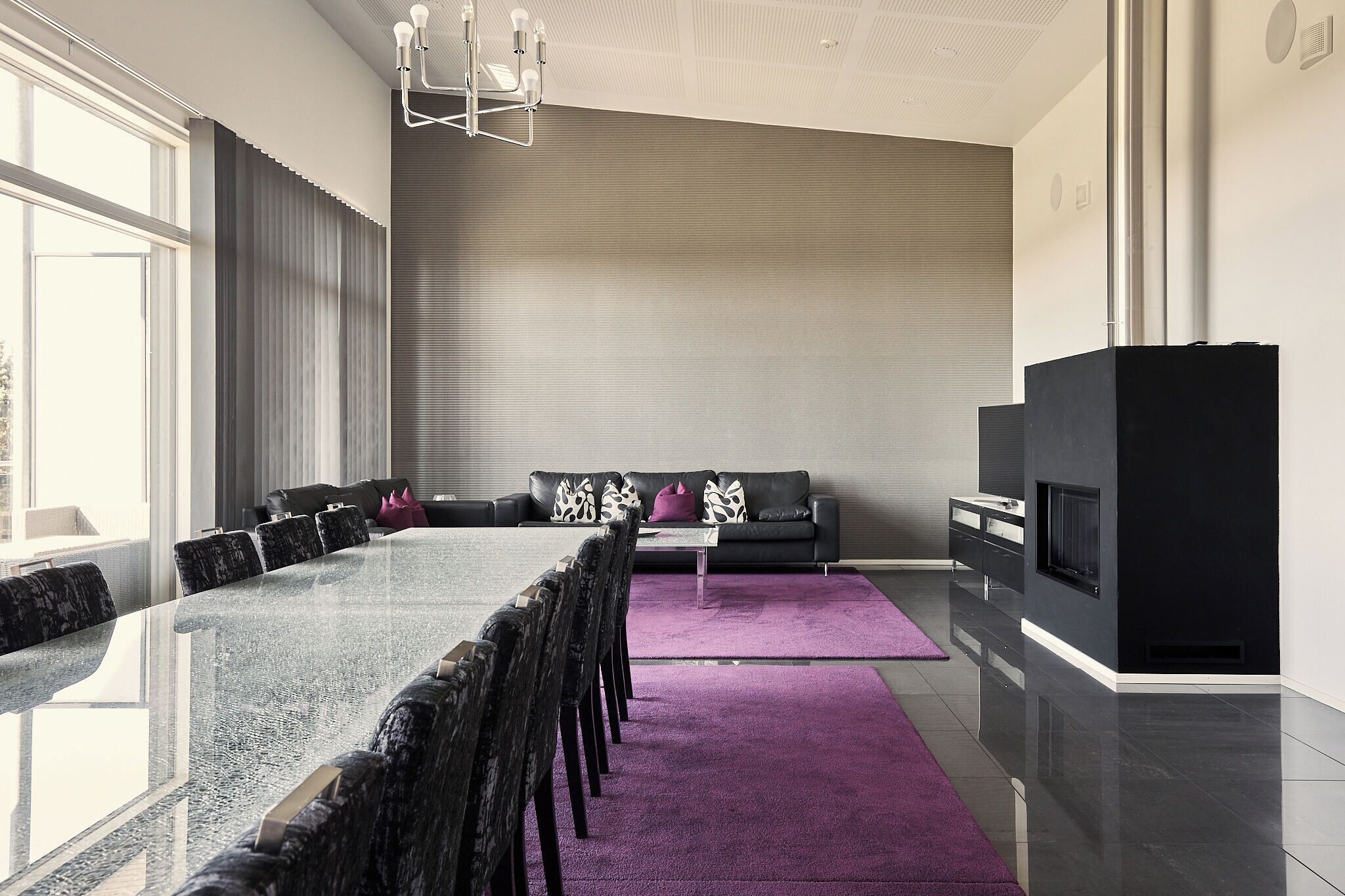 A modern living room with a glass dining table, black chairs, a black sofa with patterned cushions, a flat-screen TV, and a large window.