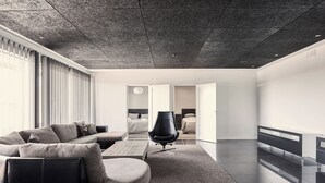 Ein modernes Wohnzimmer mit einem Ecksofa, einem schwarzen Bürostuhl und einem großen Fenster mit Jalousien.