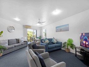 Un salon moderne avec un canapé gris, un canapé bleu et un fauteuil gris. On y trouve un ventilateur de plafond, une horloge murale et un tableau représentant une scène de plage.