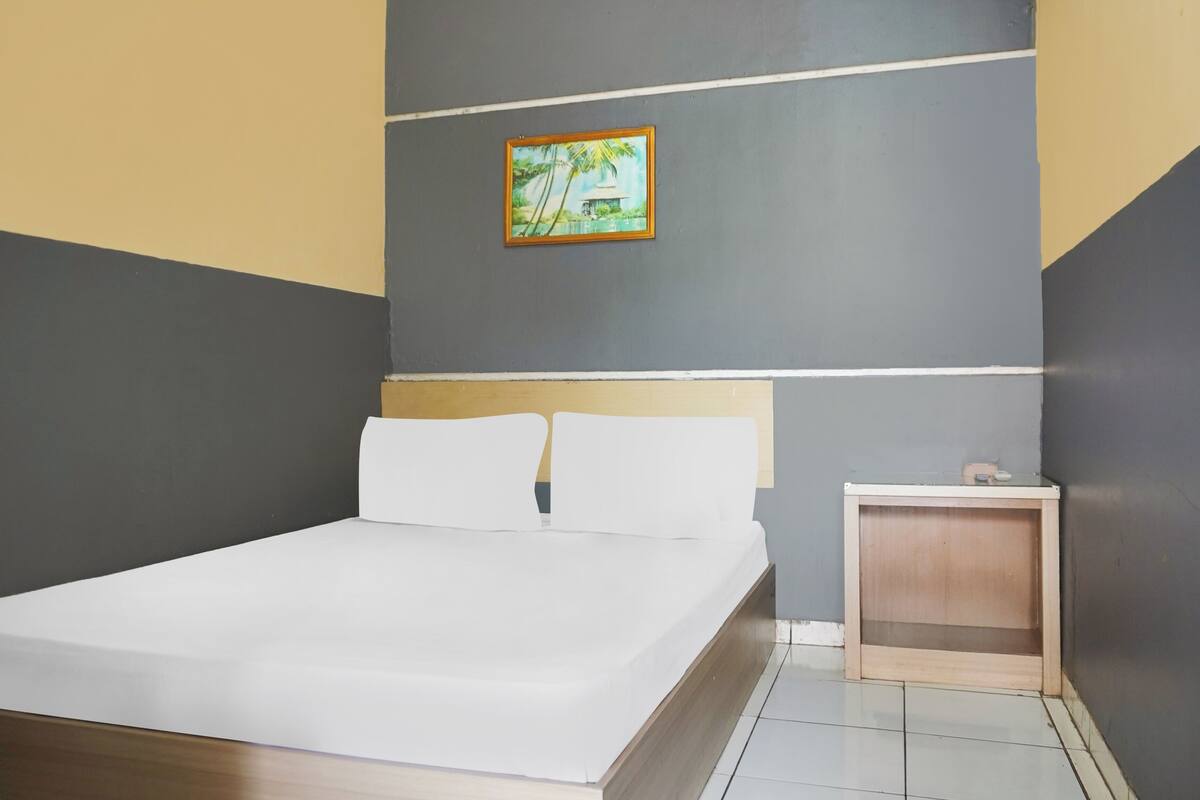 A neatly made bed in a room with a grey and yellow wall, a small wooden nightstand, and a framed picture on the wall.