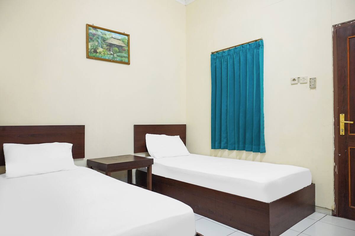 A room with two single beds, a small bedside table, a wall-mounted painting, a blue curtain, and a wooden door.
