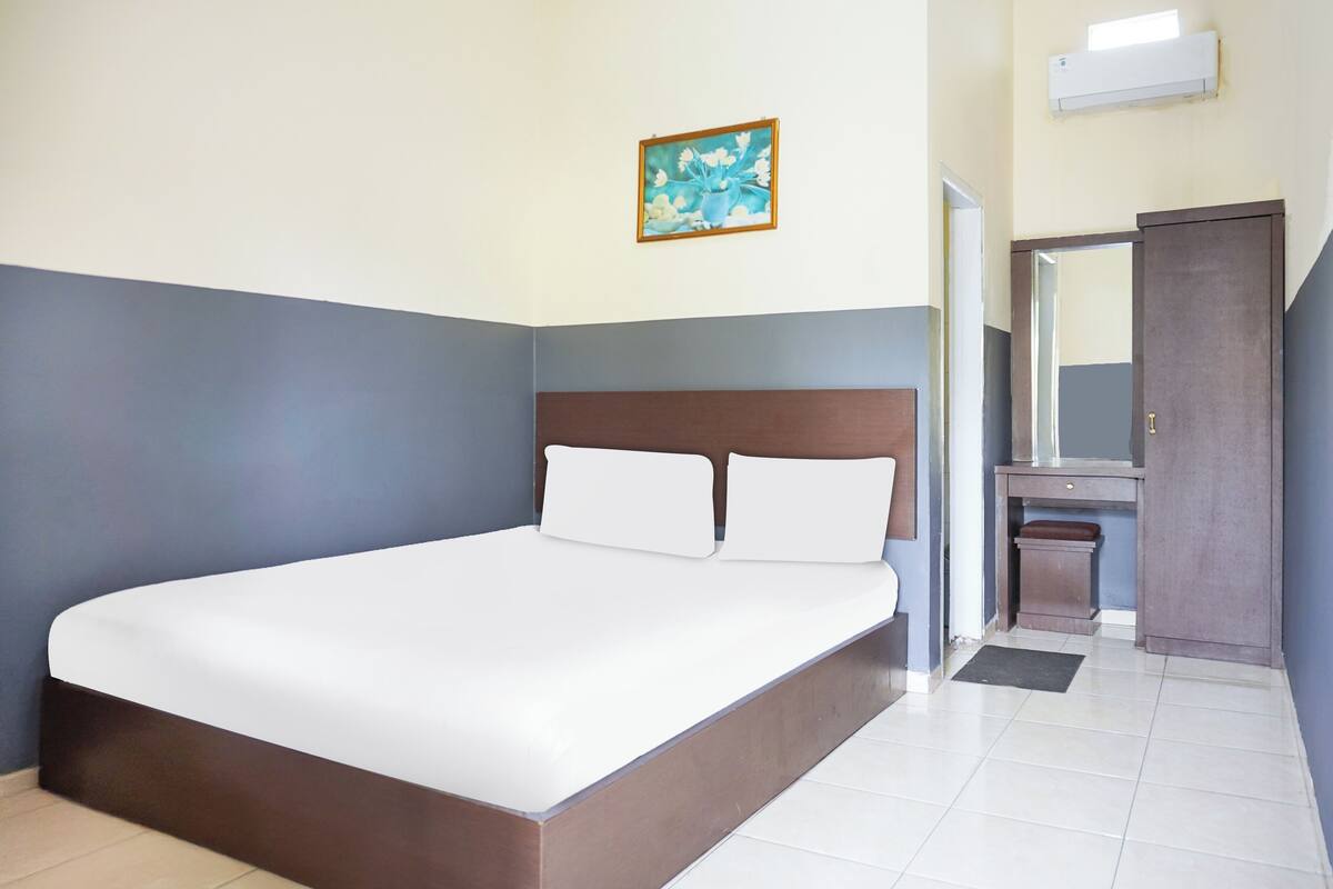 A neatly made bed in a hotel room with a headboard, a small bedside table, a mirror, and a framed picture on the wall.