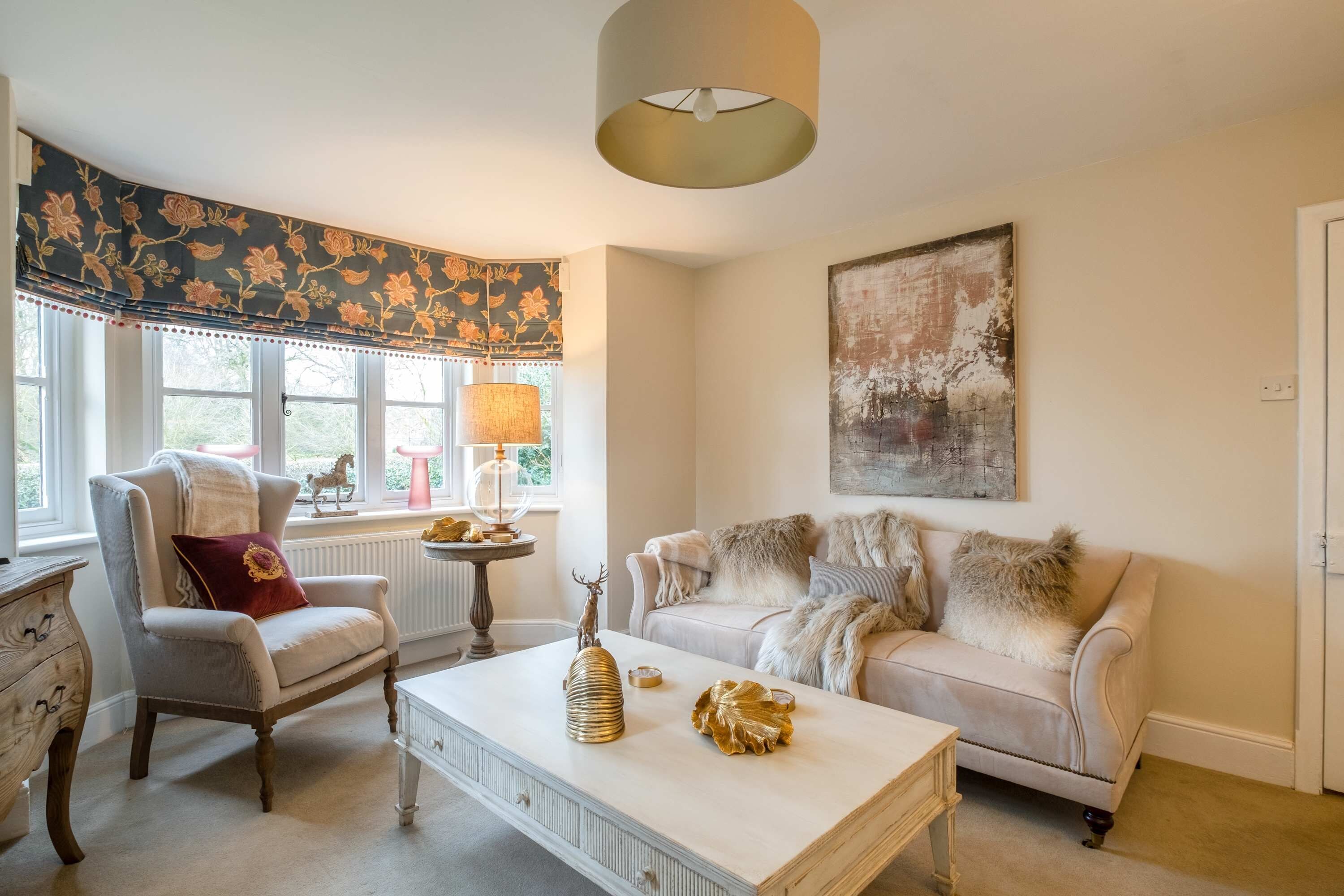 A living room with a white armchair, a beige sofa, a white coffee table, and a large window with a floral valance.