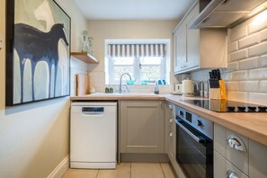 A modern kitchen with a dishwasher, oven, and a window with a striped valance. - Park Cottage (Glaston)