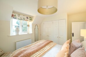 A bedroom with a large bed, a window with a view of greenery, a white wardrobe, and a ceiling light fixture. - Park Cottage (Glaston)