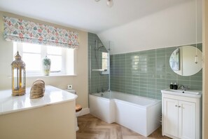 A bathroom with a bathtub, a shower, a toilet, a sink, and a window with a floral curtain. - Park Cottage (Glaston)