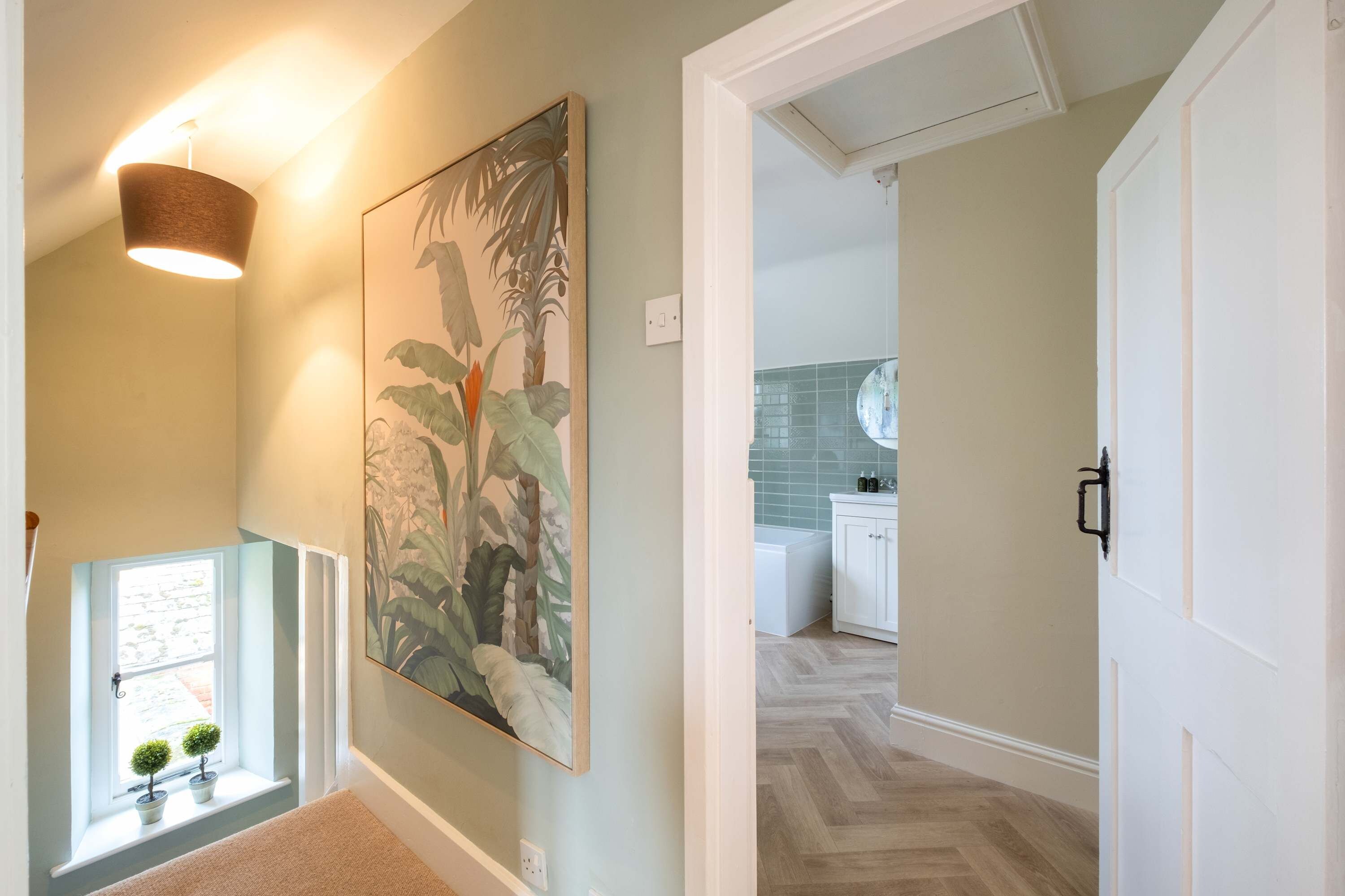 Un couloir orné d’une grande estampe représentant des motifs botaniques, un luminaire suspendu et une vue sur un espace cuisine doté d’un parquet en chevrons.