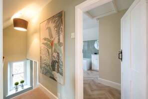 A hallway with a large botanical print on the wall, a hanging light fixture, and a view into a kitchen area with a herringbone floor. - Park Cottage (Glaston)