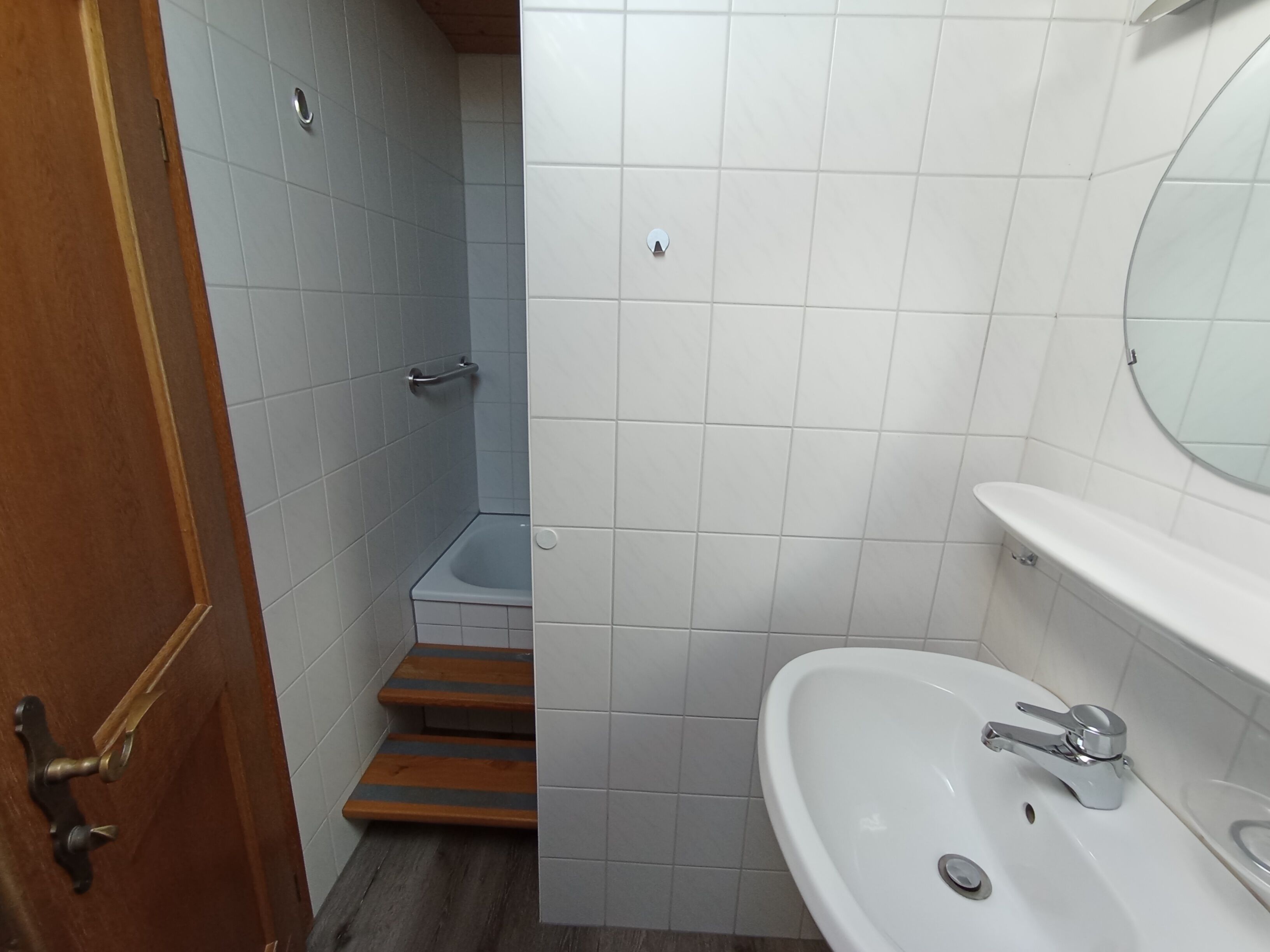 A bathroom with a wooden door, a small shower stall, and a white sink with a mirror above it.