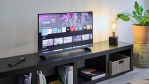 Moderni olohuone, jossa on litteänäyttöinen televisio, joka näyttää suoratoistopalvelun käyttöliittymän. Lisäksi huoneessa on kaukosäädin, ruukkukasvi ja lamppu tummanvärisellä puukonsolilla.