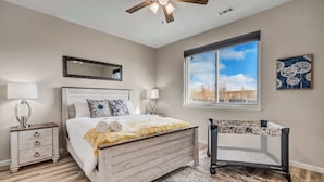 A bedroom with a wooden bed, a nightstand, a crib, a ceiling fan, a large window, and a painting on the wall.