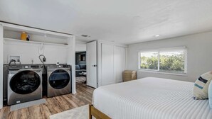A bedroom with a bed, a window with blinds, a washing machine, and a door leading to another room. - Riverside Oasis! Home w/ Tiki Hut & Firepit (Bradenton)