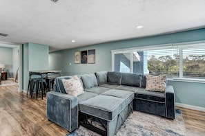 A modern living room with a sectional sofa, a dining area with a table and chairs, and a large window with blinds. - Riverside Oasis! Home w/ Tiki Hut & Firepit (Bradenton)