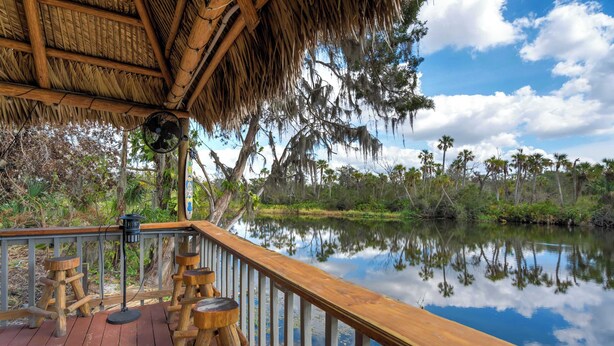 A wooden deck with a thatched roof, overlooking a calm body of water and dense vegetation. - Riverside Oasis! Home w/ Tiki Hut & Firepit (Bradenton)