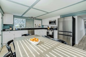 A modern kitchen with white cabinets, a stainless steel refrigerator, and a dining table with a bowl of fruit. - Riverside Oasis! Home w/ Tiki Hut & Firepit (Bradenton)