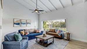 A living room with a blue sofa, a striped sofa, a coffee table, and a ceiling fan. - Riverside Oasis! Home w/ Tiki Hut & Firepit (Bradenton)