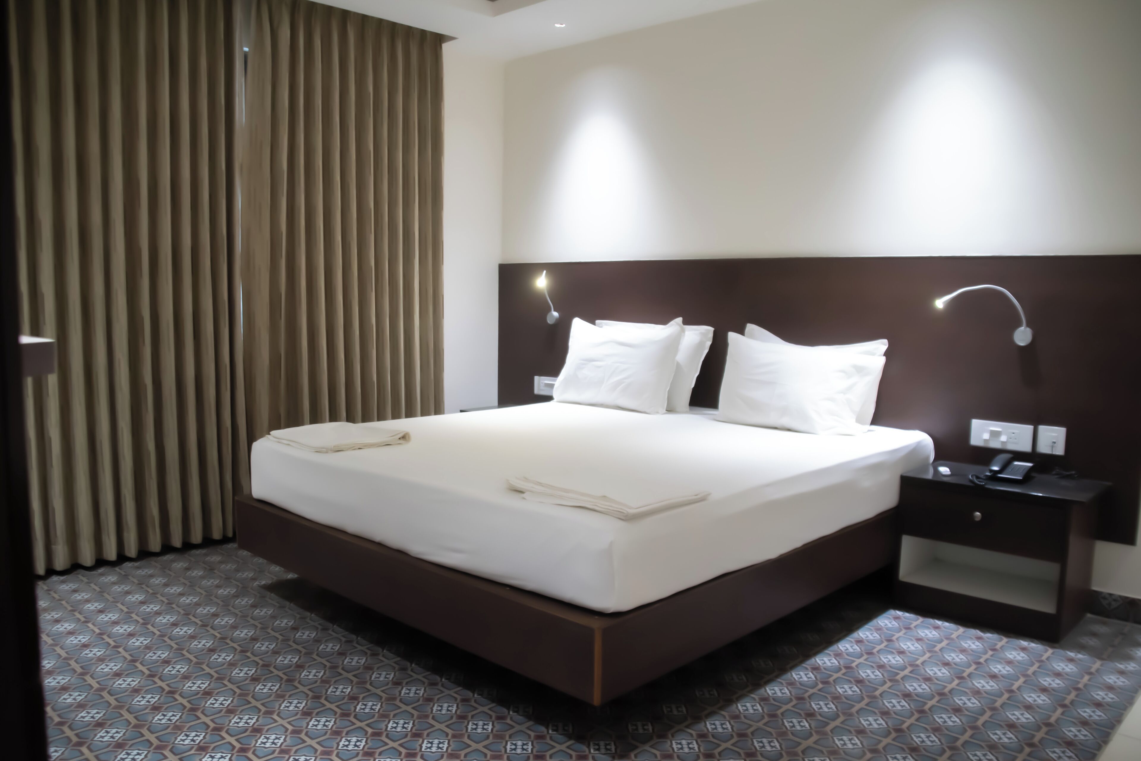 A hotel room with a large bed, two bedside tables, a wall-mounted lamp, and a patterned floor.
