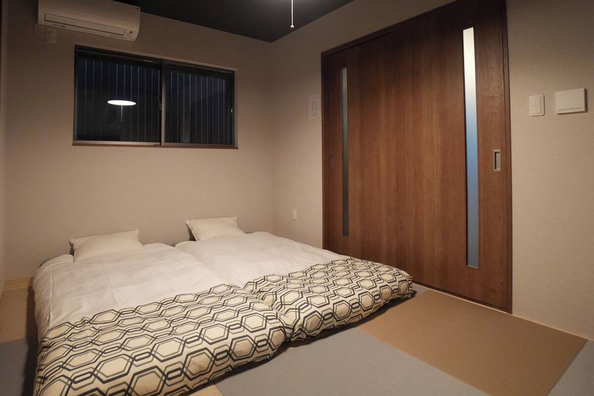 A bedroom with a bed, a window with blinds, a wooden wardrobe, and a wall-mounted air conditioner.