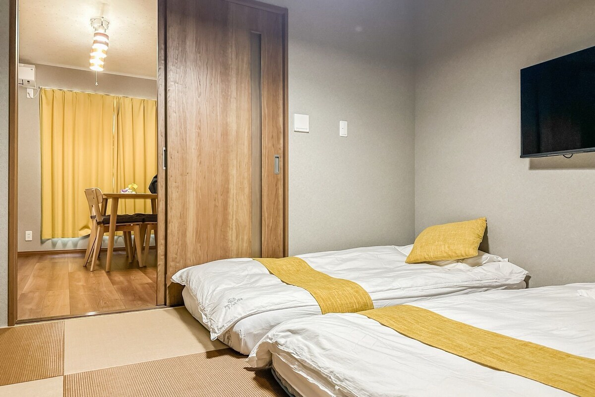 Quarto de hotel com duas camas, guarda-roupa de madeira, uma mesa pequena com cadeiras e televisão na parede.