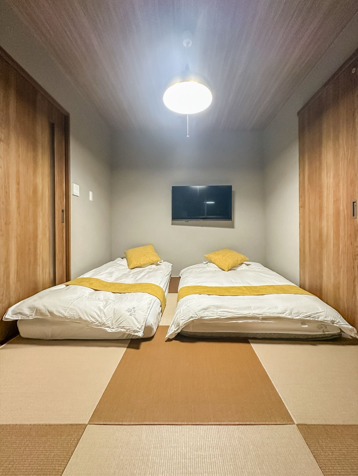 A room with two beds, a ceiling light, and a flat-screen TV on the wall.