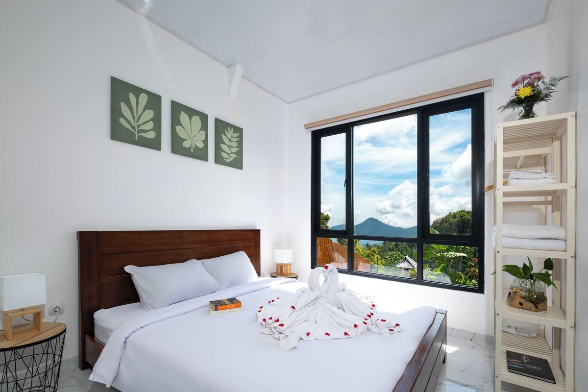 A hotel room with a large bed, a bedside table with a lamp, a window with a view of mountains, and a shelf with towels and a plant.