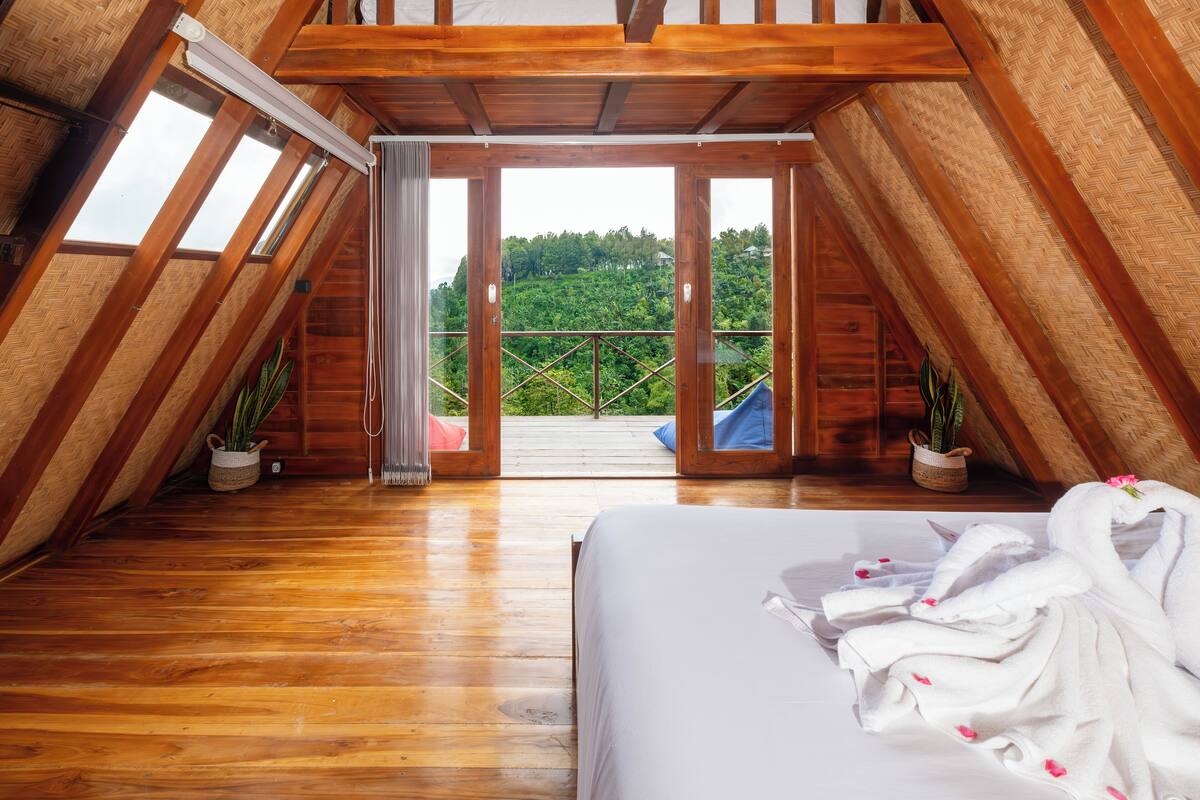 A bedroom with a wooden floor, a bed, and a view of a balcony with plants.