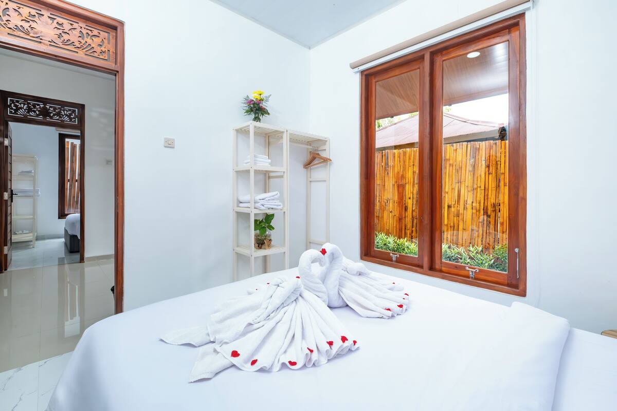 A hotel room with a bed, a wooden door, a window with a view of a bamboo fence, and a shelf with towels and a small plant.