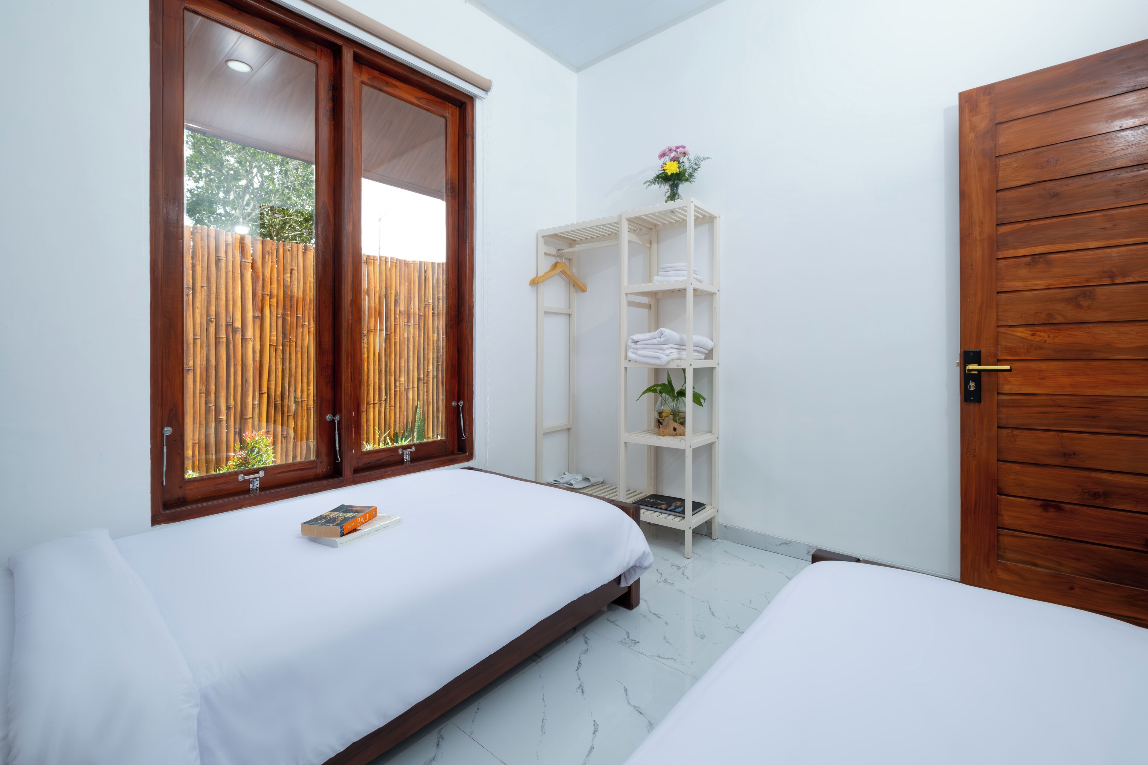 A bedroom with two beds, a wooden wardrobe, a white shelving unit, and a window with a bamboo fence view.