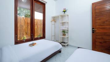 A bedroom with two beds, a wooden wardrobe, a white shelving unit, and a window with a bamboo fence view.