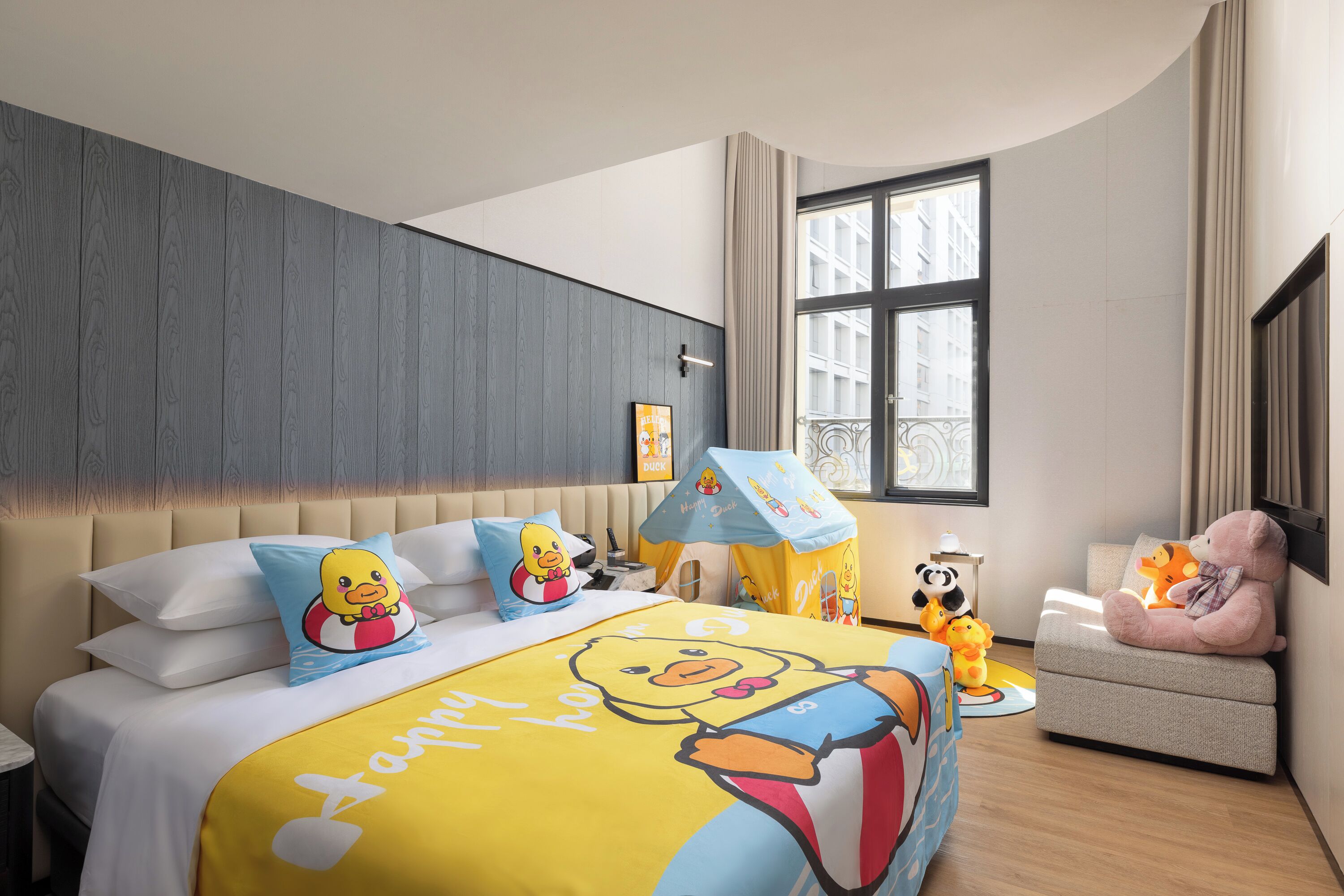 A hotel room with a bed featuring cartoon character bedding, a small play area with a tent, and a window with a view of buildings.