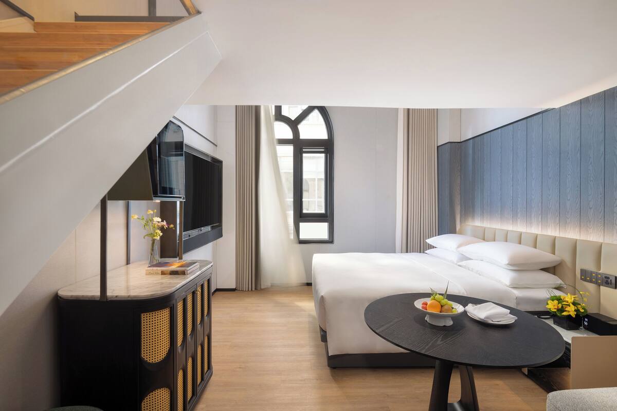 A modern hotel room with a bed, a TV, a dining table with fruit, and a view of the city.