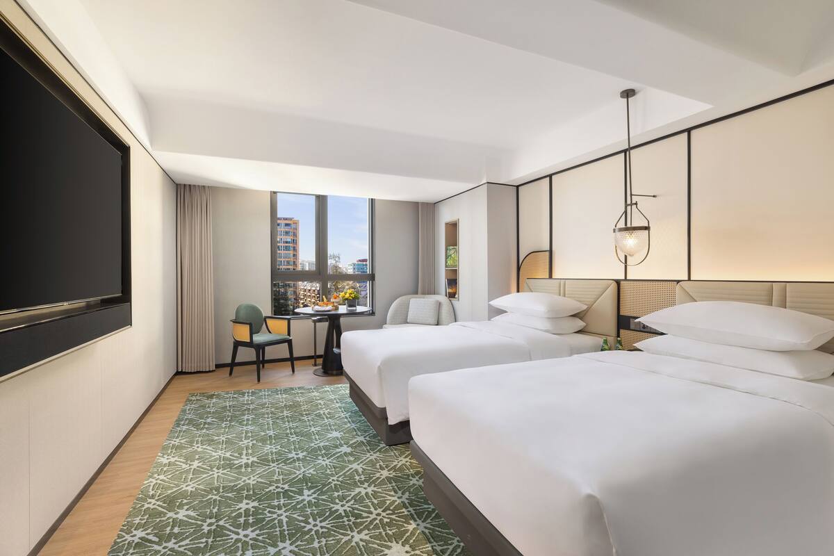A hotel room with two beds, a desk, a chair, a large window, and a TV.