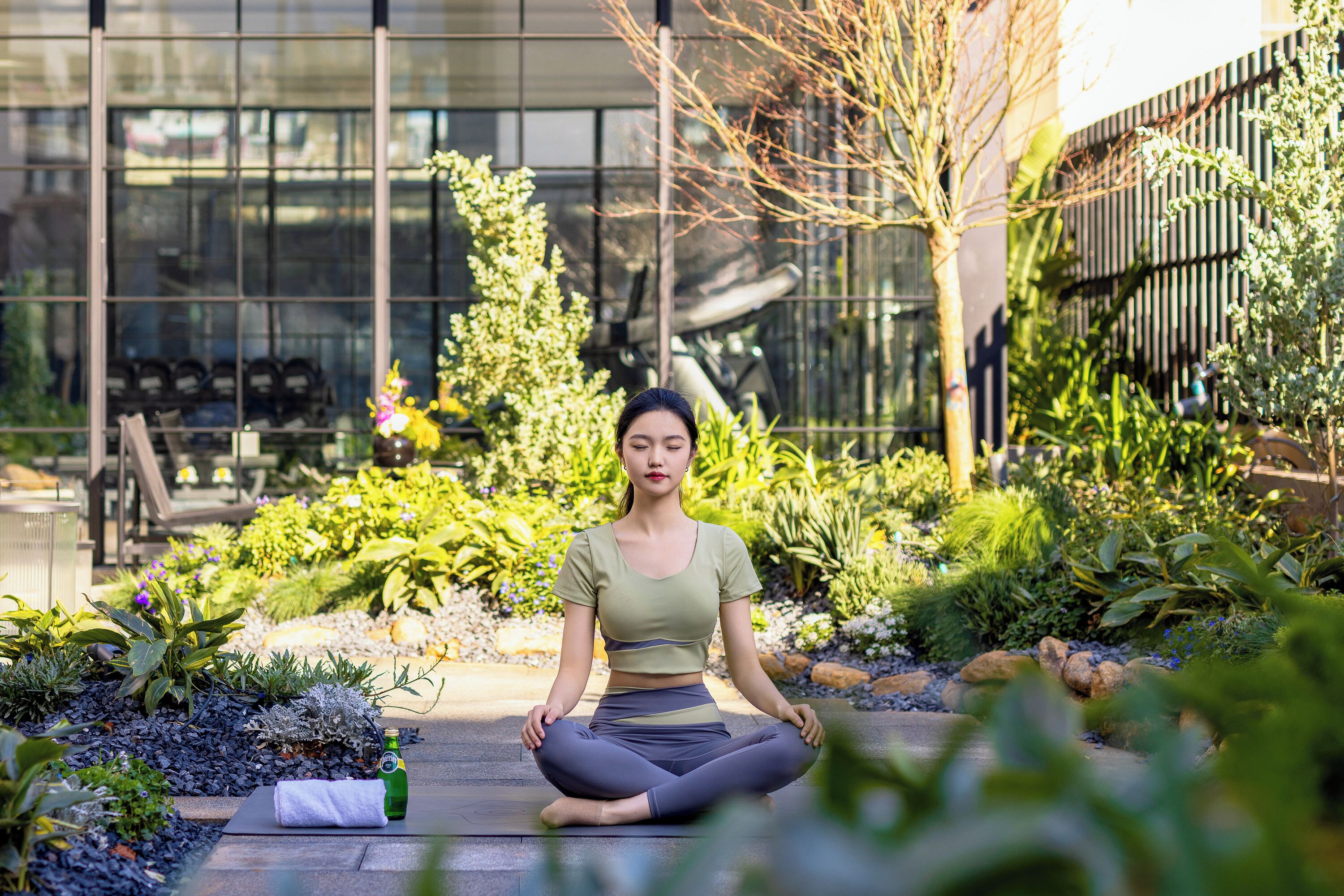 A person sitting cross-legged on a stone pathway in a garden with various plants and a building in the background.