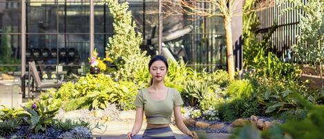 A person sitting cross-legged on a stone pathway in a garden with various plants and a building in the background.